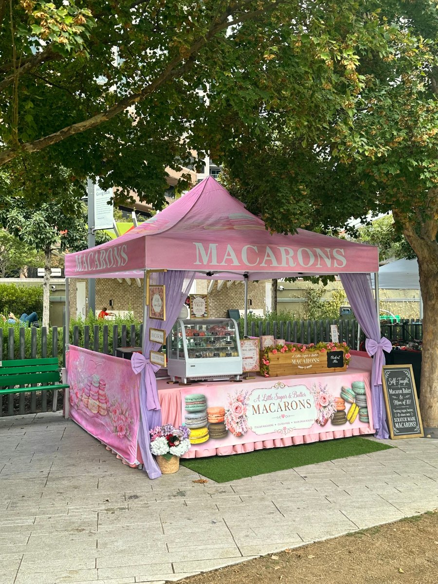 A Little Sugar & Butter Macarons booth with a pink and lavender tent, display case filled with colorful handmade macarons, and signs advertising gluten-free macarons and other homemade baked goods.